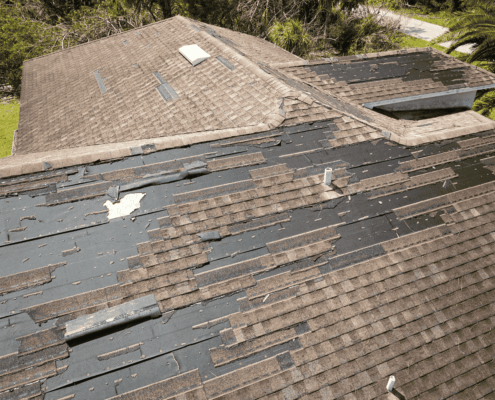The image displays significant storm damage to a roof, characterized by missing and peeling shingles, exposing the underlying roofing materials.